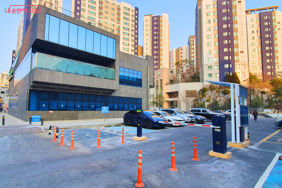 독서당 인문아카데미센터 건물 바로 옆에 위치한 금호스포츠센터 노외주차장의 모습이다. 주차장 입구에는 무인 정산 시스템과 결합된 차량 차단기(붐 게이트)가 설치되어 있으며, 주차장 내부에는 여러 대의 승용차가 주차되어 있다. 주차장 너머로는 센터 건물의 유리 외벽과 대규모 고층 아파트 단지가 배경으로 보이며, 장애인 전용 주차 구역을 알리는 파란색 표지판과 안전을 위한 주황색 탄력봉이 세워져 있다. &copy;양정화