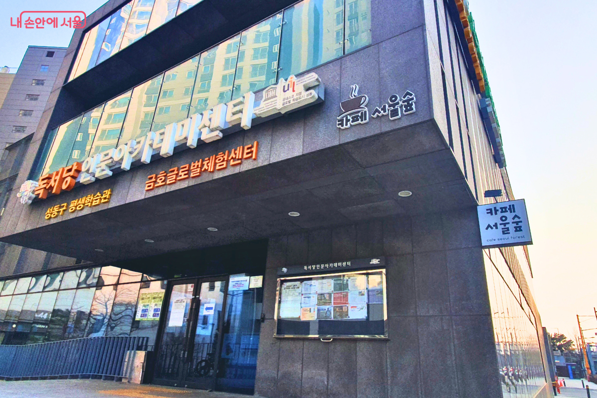 센터의 주 출입구와 외벽을 근접 촬영한 사진이다. 입구 상단에는 '독서당 인문아카데미센터' 명칭과 함께 유네스코 글로벌 학습도시 로고가 부착되어 있다. 우측 외벽에는 '카페 서울숲'의 로고와 돌출 간판이 설치되어 있어 센터 내부에 카페가 운영되고 있음을 알려준다. 입구 옆 게시판에는 다양한 홍보물이 부착되어 있다. &copy;양정화