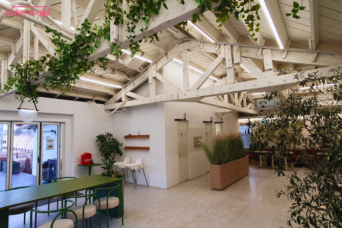 하얀색 서까래가 노출된 높은 층고의 카페 어울림 내부다. 곳곳에 배치된 초록색 식물과 나무들이 쾌적한 온실 같은 분위기를 자아낸다.