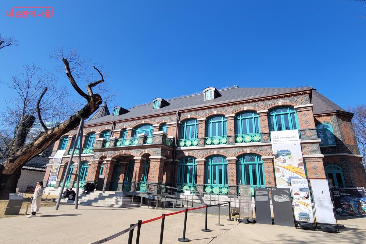 서울 덕수궁 내 돈덕전 건물 외관 모습으로, 붉은 벽돌 2층 건물과 청록색 창틀, 전시 안내 배너가 보이고 앞에는 관람객과 통제 라인이 설치되어 있다.