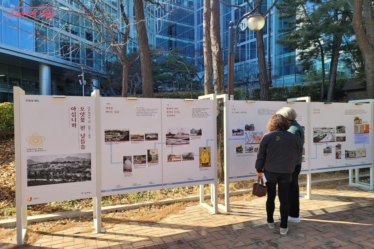 정동 거리의 역사 안내 전시판을 시민들이 읽고 있는 모습으로, 대한제국 관련 사진과 설명이 담겨 있다.