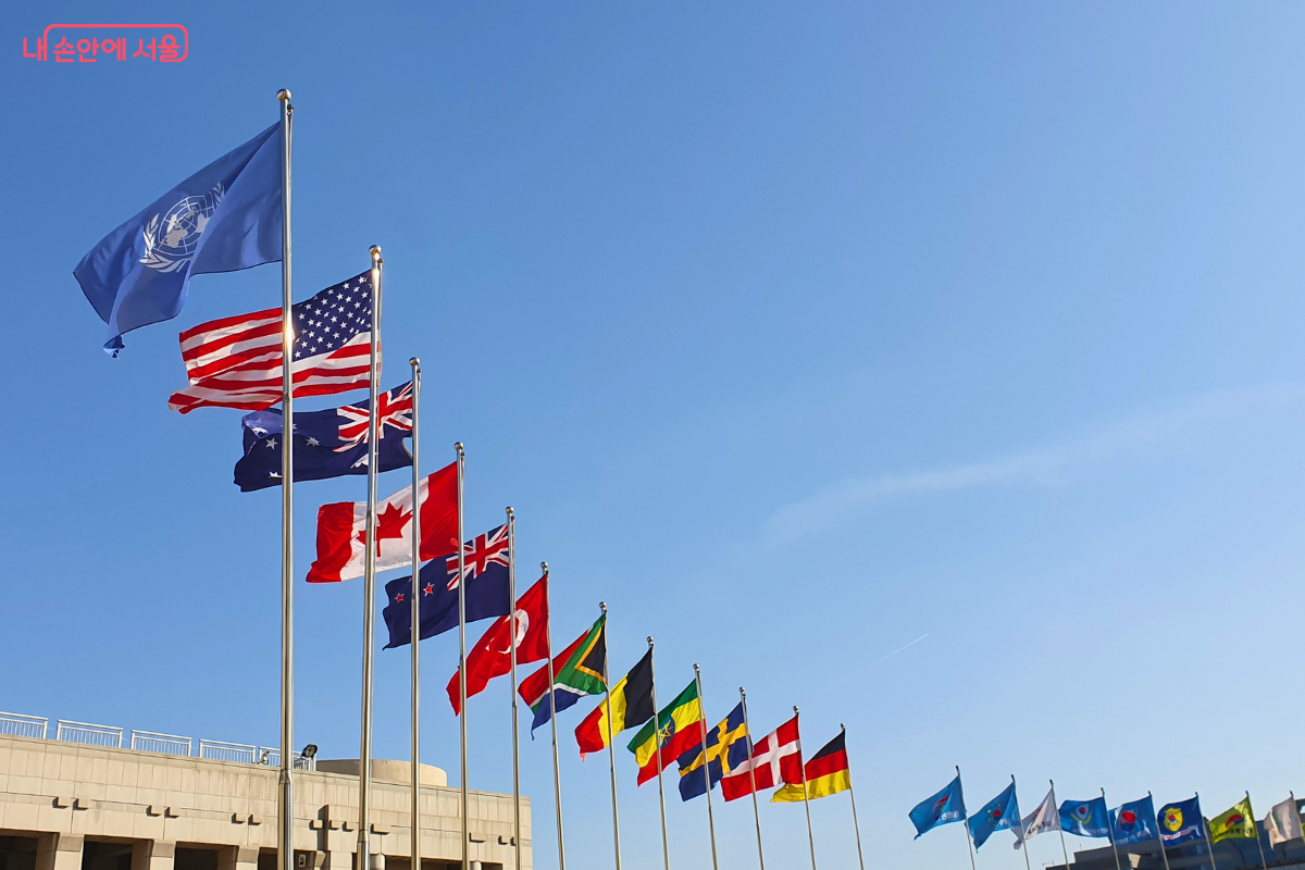 전쟁기념관 광장에 나란히 게양된 6·25전쟁 참전국들의 국기 모습이다. 가장 앞쪽에 UN기와 미국 국기가 높게 배치되어 있으며, 그 뒤로 호주, 캐나다, 뉴질랜드 등 참전국들의 국기가 푸른 하늘을 배경으로 일렬로 펄럭이고 있다. 하단에는 본관 건물의 일부가 보인다.