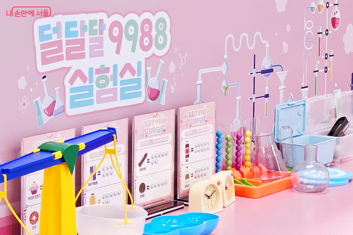 키즈라운지의 덜달달 구구팔팔 실험실 사진. 덜달달 구구팔팔 실험실이라는 제목과 여러 실험도구 일러스트의 분홍색 배경 공간이 있다. 그 안에는 각양각색의 실험도구 장난감과 음식의 설탕 함량이 적힌 팻말이 비치되어 있다.