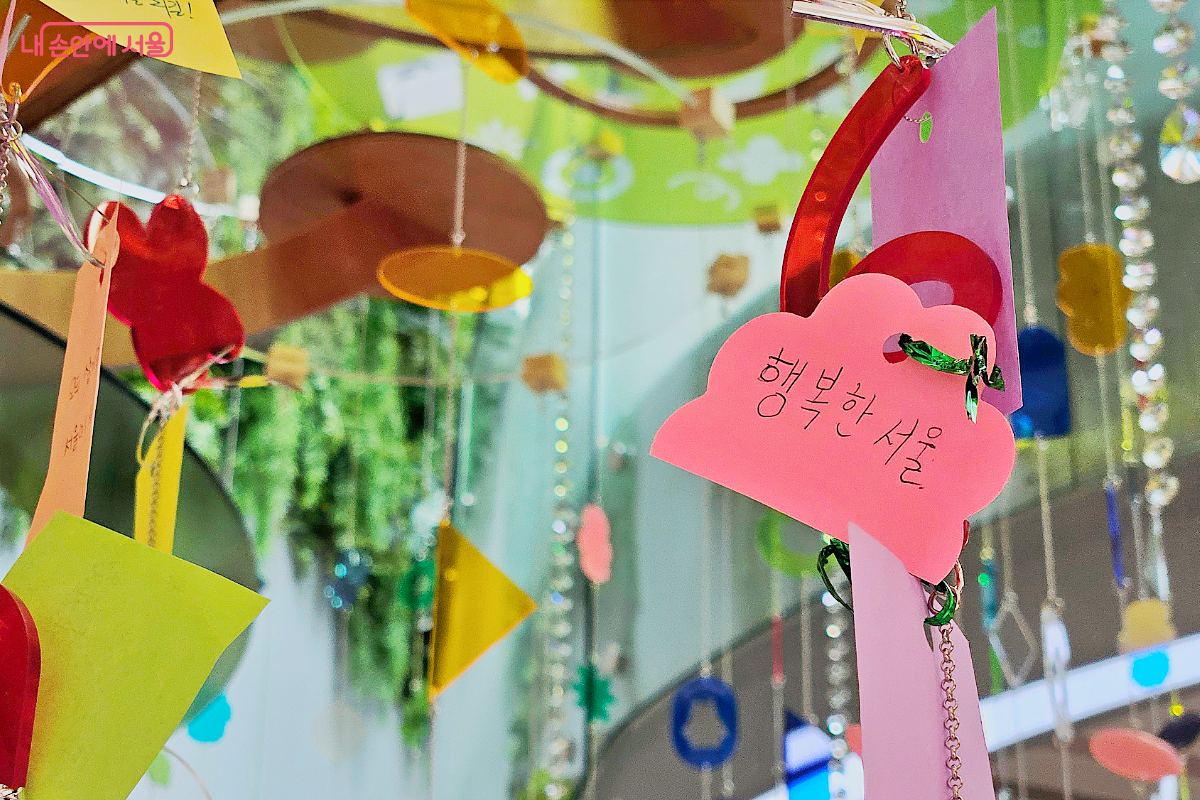 서울갤러리 소망나무 사진. 알록달록한 아크릴 오브제와 비즈가 주렁주렁 매달려 있다. 행복한 서울이라고 적혀 있는 분홍색 쪽지가 걸려 있다.