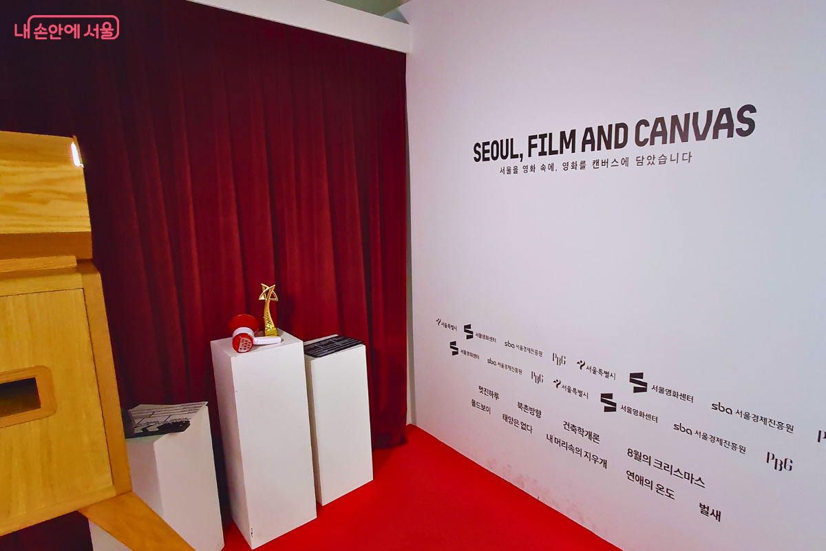 전시장 한쪽 구역에 마련된 포토존의 모습이다. 왼쪽에는 짙은 붉은색 벨벳 커튼이 드리워져 무대 분위기를 자아내고, 그 앞의 흰색 좌대 위에는 빨간색 촬영용 확성기와 황금색 트로피가 놓여 있다. 오른쪽 흰 벽면에는 전시 제목과 함께 서울특별시, 서울영화센터, 서울경제진흥원 등 이번 전시를 주최하고 후원한 관계 기관들의 로고가 공식적으로 나열되어 있다. ©양정화
