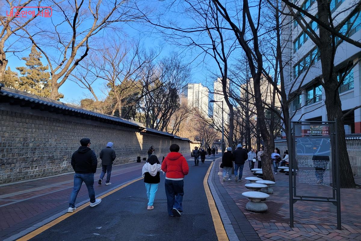 덕수궁 돌담 옆 정동길을 시민들이 걷고 있으며, 안내판과 가로수가 보이는 도심 산책로 모습.