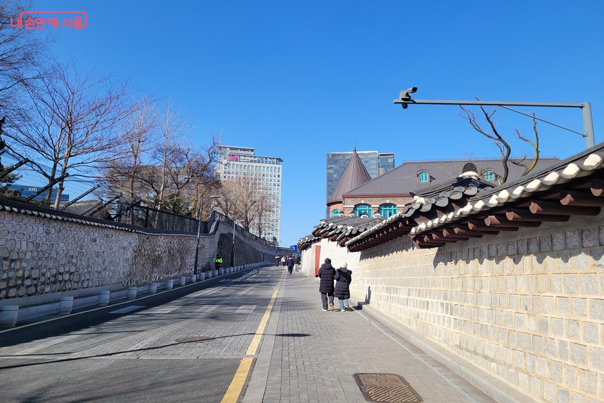 덕수궁 전통 담장을 따라 이어진 돌담길을 시민들이 걷고 있으며, 뒤편으로 도심 건물이 보이는 모습.
