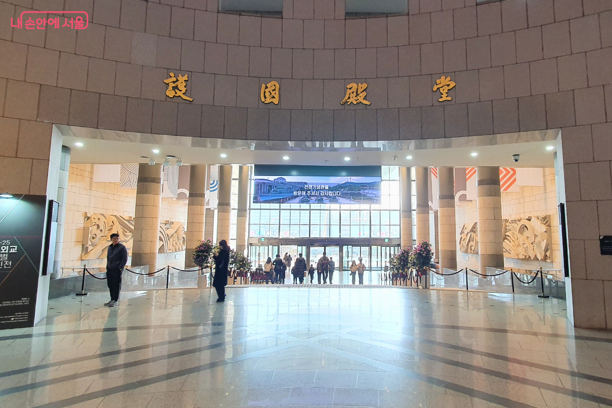 전쟁기념관 본관 내부에서 중앙 출입구 방향을 바라본 모습이다. 입구 상단 벽면에는 한자로 호국전당이라는 문구가 금색으로 새겨져 있으며, 그 아래로 대형 전광판과 외부로 나가는 유리문이 보인다. 대리석 바닥에는 기하학적인 문양이 새겨져 있고 기둥들이 질서 있게 배치되어 있다.