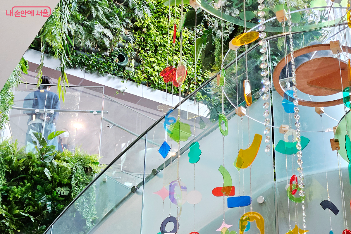 알록달록한 아크릴 오브제와 비즈가 주렁주렁 매달린 소망나무. 소망나무 너머로 서울 시청과 서울갤러리를 잇는 계단이 있다.