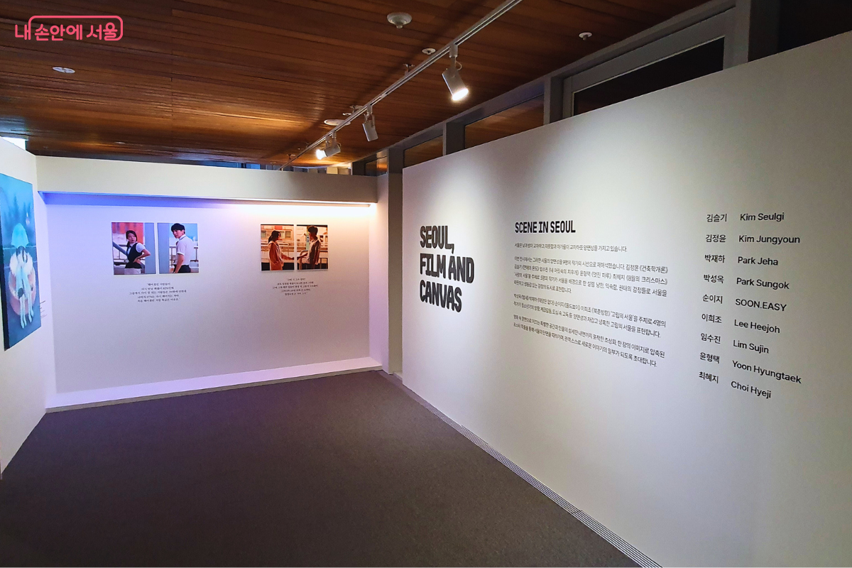 서울영화센터 4층 전시실 내부의 도입부 벽면 모습이다. 정면의 흰색 가벽에는 'SEOUL, FILM AND CANVAS'라는 전시 제목과 'SCENE IN SEOUL'이라는 문구, 그리고 이번 전시에 참여한 9명의 작가 명단이 검은색 글씨로 깔끔하게 정렬되어 있다. 왼쪽 벽면에는 영화 장면을 재해석한 파란색 톤의 회화 작품과 영화 스틸컷이 조명을 받으며 나란히 전시되어 관람객을 맞이한다. ©양정화