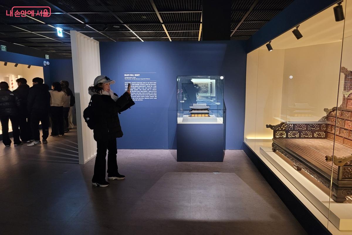 덕수궁 돈덕전 전시 내부 모습으로, 유물이 진열된 유리 전시관과 설명 패널을 관람하는 방문객이 보이는 장면.