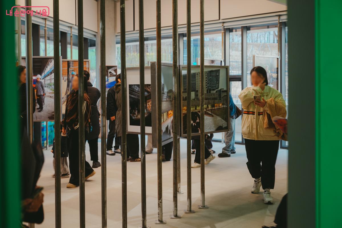 전시실 내부. 철제 기둥들 사이로 공사 현장의 모습을 담은 건축 기록 사진들이 걸려 있고, 관람객들이 이를 살펴보고 있다.
