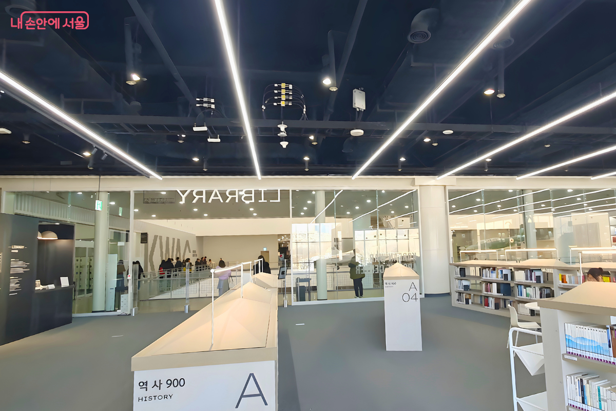 자료실 내부에서 유리 입구 방향을 바라본 시점이다. 천장은 검은색 노출형으로 되어 있고 격자 형태의 긴 LED 조명들이 달려 있다. 중앙에는 낮은 서가가 배치되어 있으며, 유리 벽 너머로 복도를 지나가는 사람들의 모습이 보인다. 좌측 벽면에는 검은색 안내 패널이 설치되어 있다.