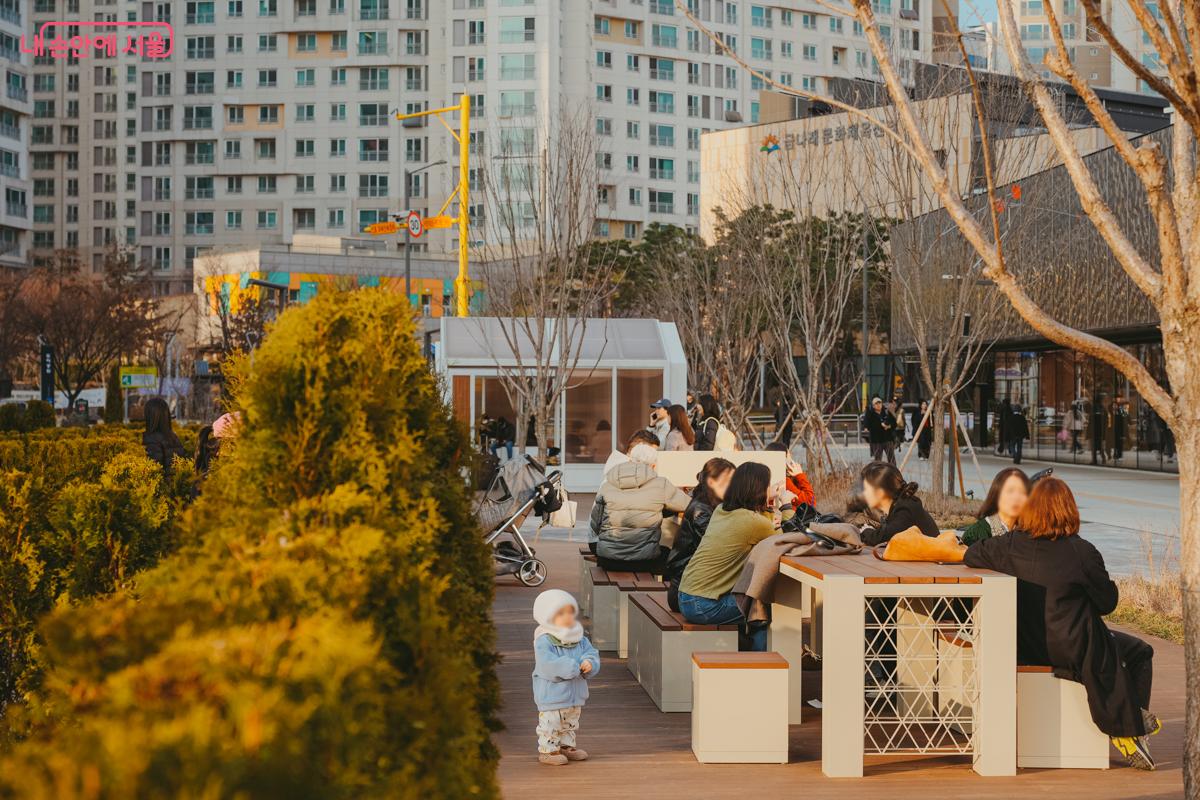 공원 야외 테이블 벤치. 따뜻한 햇살 아래 시민들이 삼삼오오 모여 앉아 담소를 나누고 있고, 곁에 털옷을 입은 어린아이가 서 있다.