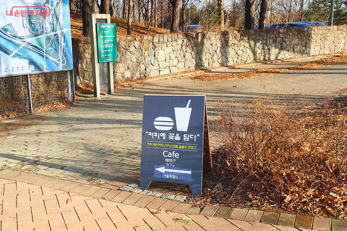 서울숲 산책로 입구에 세워진 남색 안내 입간판이다. 상단에는 햄버거와 컵 모양의 픽토그램이 그려져 있고, &lsquo;커피에 꽃을 담다&rsquo;라는 상호와 함께 커피, 베이커리, 아이스크림, 음료수, 핫도그 등의 메뉴가 한글과 외국어로 병기되어 있다. 하단에는 화살표와 함께 서울특별시 로고가 포함되어 카페의 위치를 안내한다. &copy;양정화
