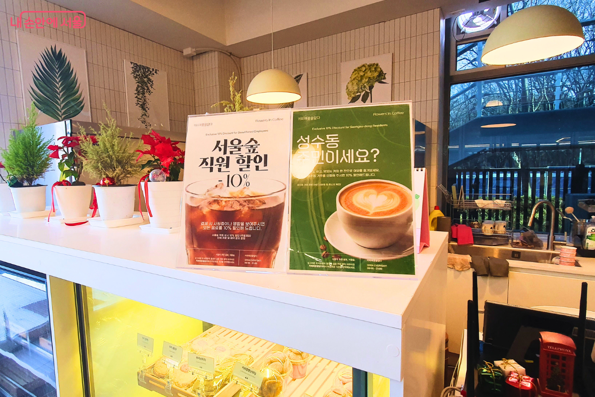 카페 내부 카운터 위에 비치된 할인 안내 포스터들이다. 왼쪽에는 &lsquo;서울숲 직원 할인 10%&rsquo;, 오른쪽에는 초록색 바탕의 &lsquo;성수동 주민이세요?&rsquo;라는 문구와 함께 10% 할인 혜택을 알리는 안내판이 나란히 세워져 있다. 안내판 뒤로는 실내를 장식한 화분들과 메뉴 진열장이 일부 보인다. &copy;양정화