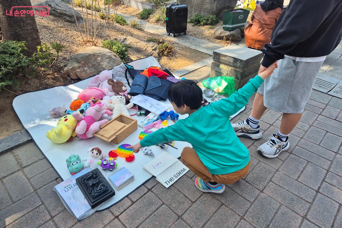 아이가 아빠의 손을 잡고 장터의 장난감을 가리키며, 사달라고 조르고 있다. ©마가은