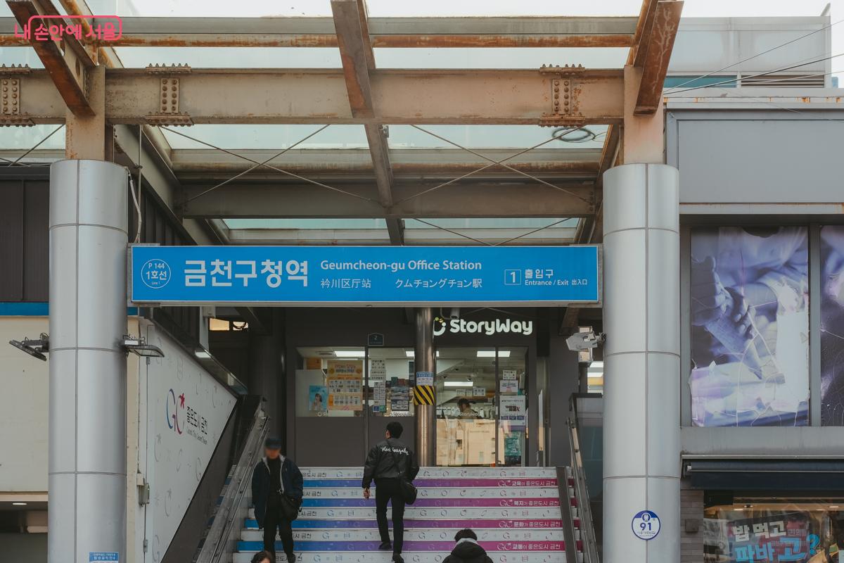 지하철 1호선 금천구청역 1번 출구 전경. 파란색 역 간판 아래로 시민들이 계단을 오르내리고 있다.