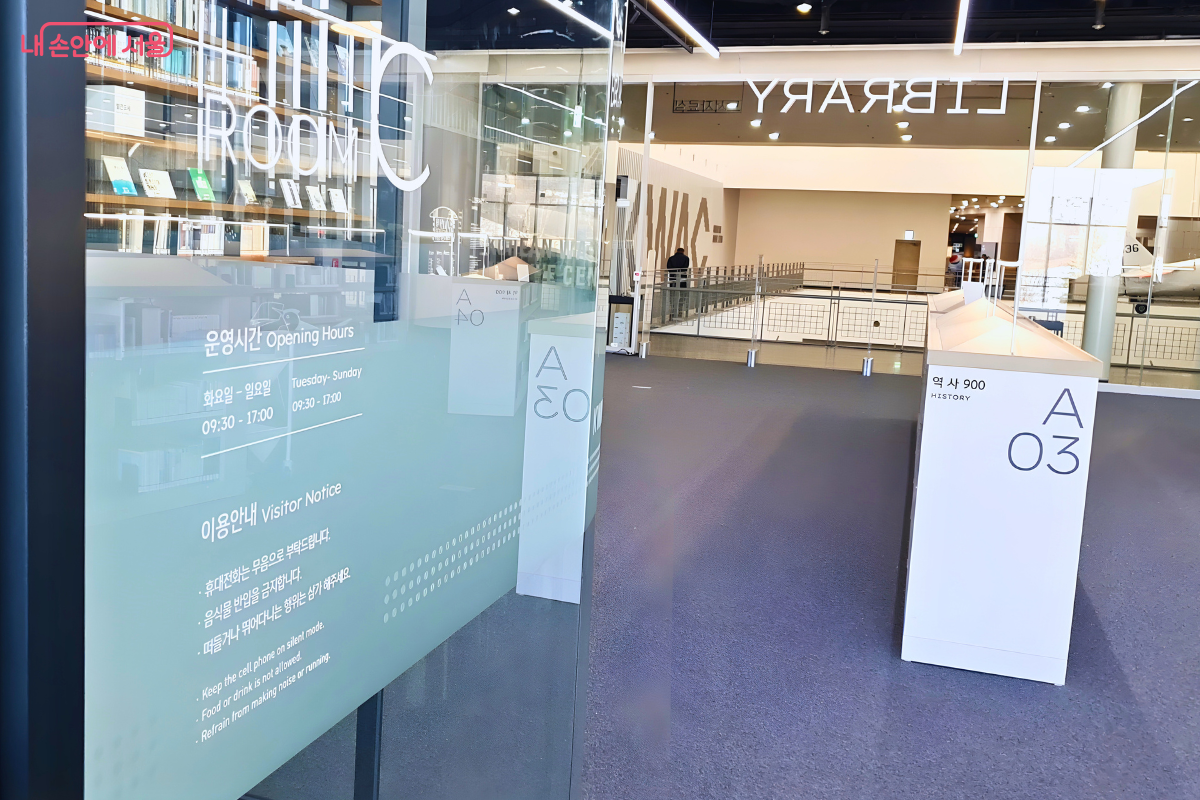 투명한 유리 벽으로 분리된 세미나실의 외관이다. 유리 벽면에는 '운영시간 Opening Hours 화요일-일요일 09:30-17:00'과 '이용안내 Visitor Notice' 문구가 흰색 시트지로 부착되어 있다. 유리 벽에는 내부 서가와 반대편 복도의 모습이 반사되어 보인다.