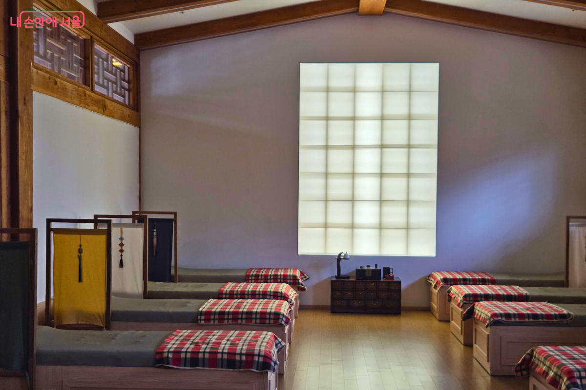 전신 마사지 체험을 위한 아늑한 실내 공간이다. 낮은 목제 침대들이 일정한 간격으로 놓여 있으며, 각 침대 위에는 빨간색과 검은색이 섞인 격자무늬 담요가 덮여 있다. 침대 머리맡에는 노란색과 흰색 천이 드리워진 전통 창호 느낌의 파티션이 설치되어 있고, 정면 벽에는 큰 사각 격자 형태의 불투명 채광창이 있다.