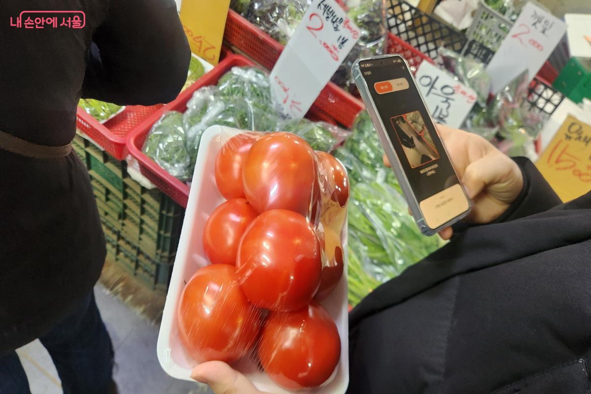 한 고객이 구매할 토마토를 들고 스마트폰에서 디지털온누리 앱을 실행해 QR코드 결제를 준비하고 있다. 휴대전화 화면에는 결제를 위한 QR 기능이 표시돼 있다.휴대전화 화면은 QR을 인식하기 위한 카메라 모드로 설정돼 있다.