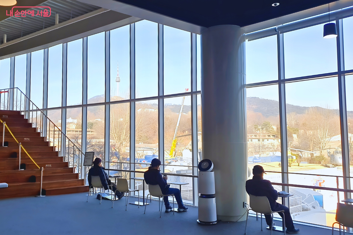 격자무늬의 대형 통창을 통해 외부 전경이 시원하게 보이는 열람실 내부이다. 창밖 멀리 산 정상에 N서울타워가 보이며, 바로 앞 야외에는 헬리콥터와 항공기 전시물들이 내려다보인다. 실내 창가에는 1인용 테이블과 의자들이 일렬로 배치되어 있으며, 세 명의 이용자가 앉아 있다. 좌측에는 나무 계단이, 중앙에는 공기청정기가 놓여 있다.