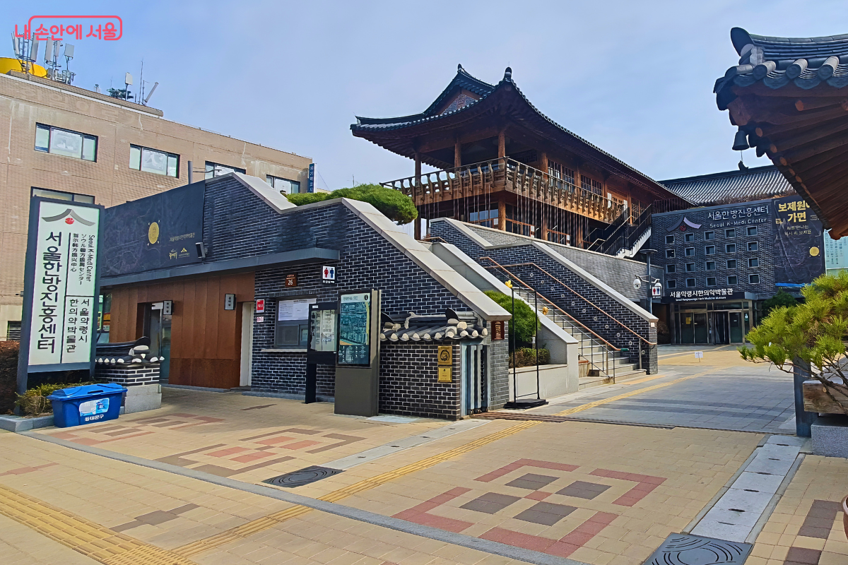 서울한방진흥센터의 외부 전경이다. 현대적인 짙은 회색 전벽돌 건물 위에 웅장한 기와 누각이 올려진 독특한 건축 구조를 보여준다. 좌측에는 '서울한방진흥센터'와 '서울약령시 한의약박물관' 명칭이 세로로 적힌 대형 입간판이 서 있다. 중앙에는 상층부로 이어지는 야외 돌계단이 길게 설치되어 있다.