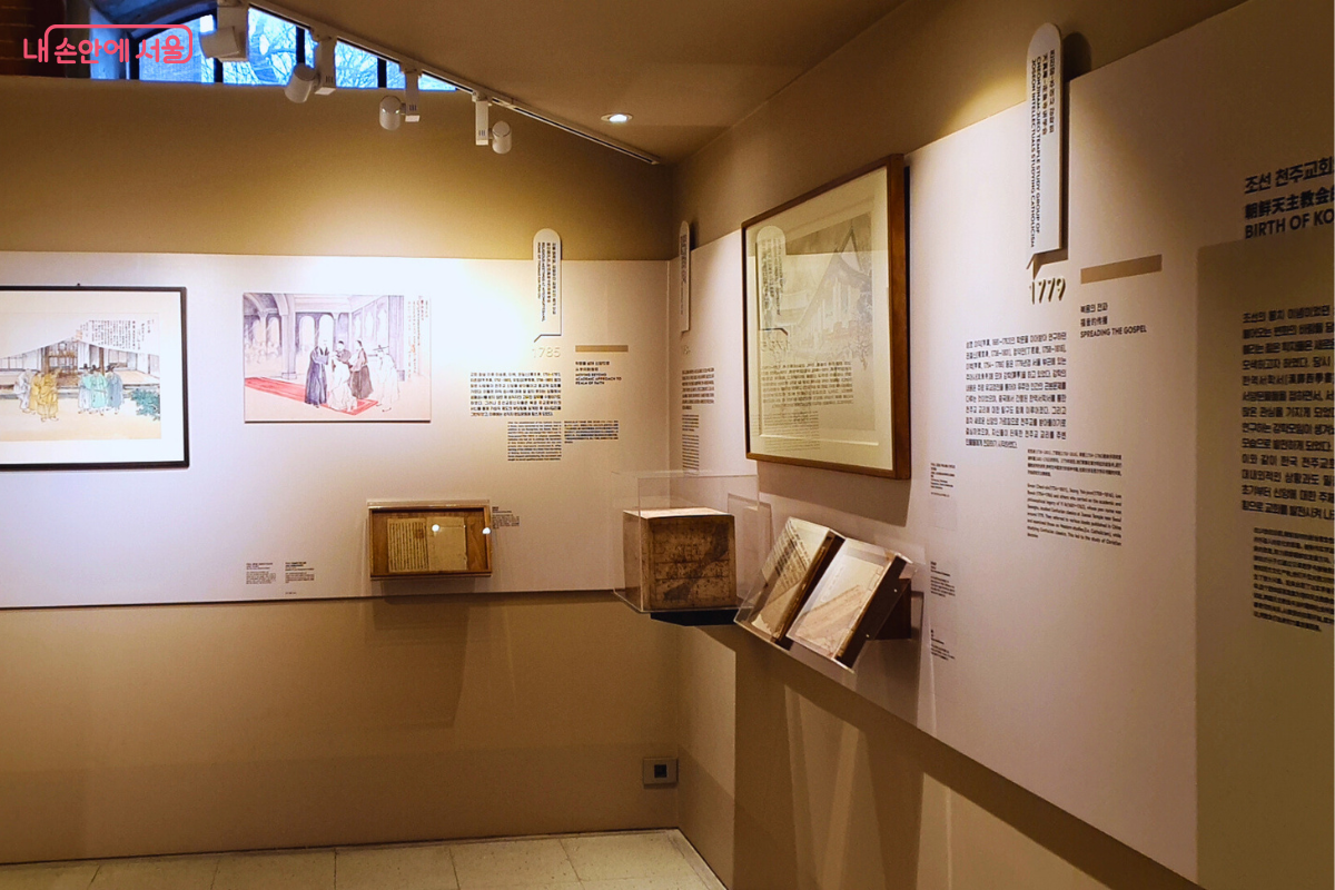 '조선 천주교회의 태동'을 주제로 한 전시 공간이다. 베이지색 벽면에 여러 점의 역사적 그림 액자와 '1779' 등 연도가 적힌 설명판이 부착되어 있다. 중앙 유리 진열장 안에는 오래된 서적이 펼쳐진 채 전시되어 있다. &copy;양정화