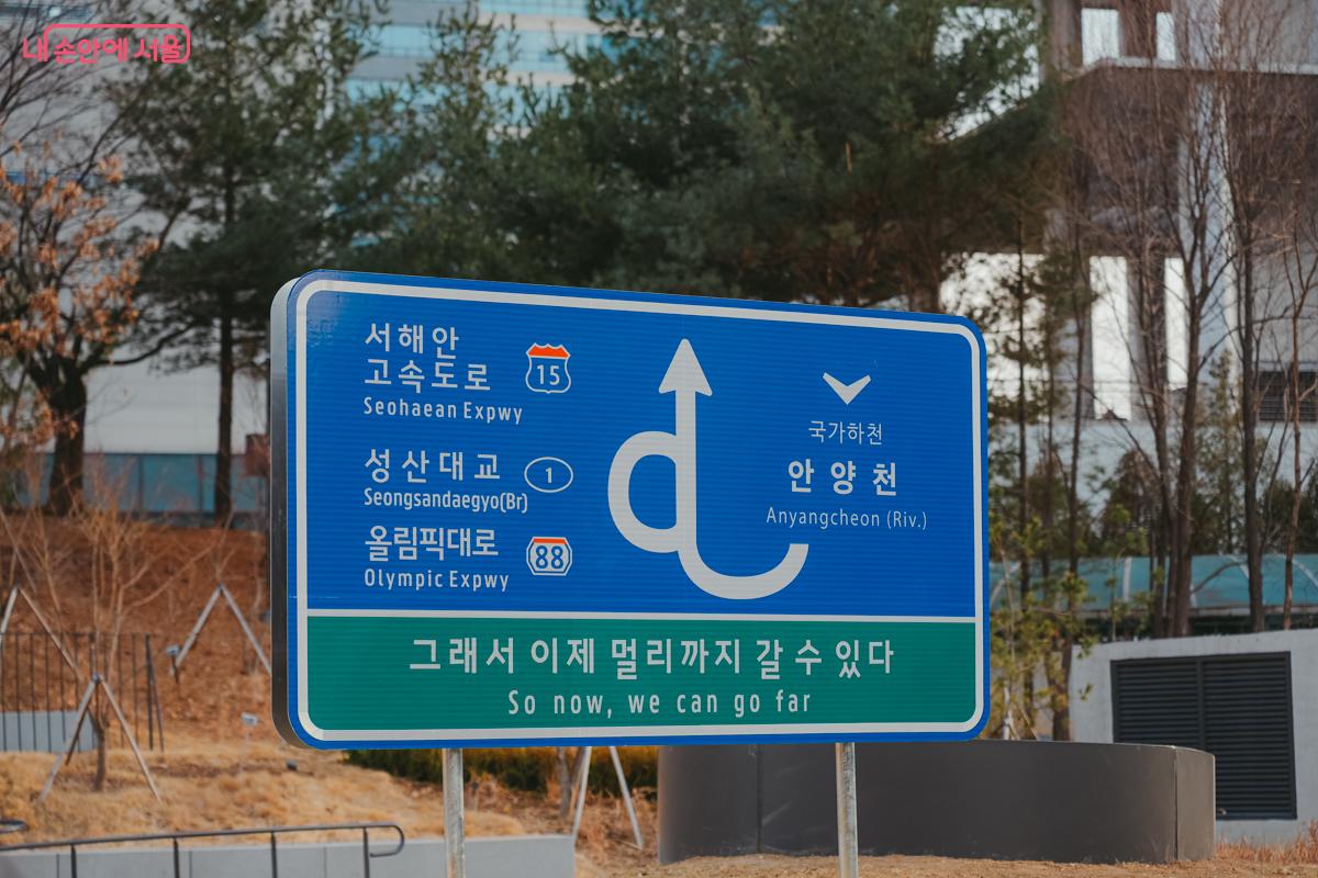 파란색 고속도로 표지판 형태의 설치 작품. 복잡한 화살표 기호 아래 '그래서 이제 멀리까지 갈 수 있다'라는 글귀가 적혀 있다.