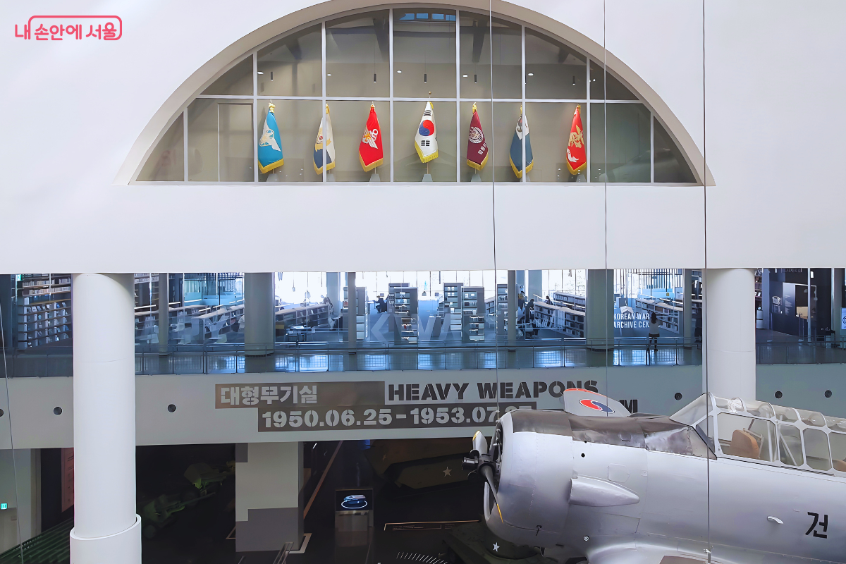 대형 아치형 구조물 너머로 보이는 도서관 내부와 하층 전시실의 모습이다. 아치 상단에는 태극기를 포함한 7개의 깃발이 나란히 꽂혀 있다. 2층에는 낮은 서가들이 배치된 'KWAC' 라이브러리 공간이 보이며, 그 아래 1층에는 '대형무기실'이라고 적힌 전시실이 있다. 우측 하단에는 실제 크기의 은색 비행기 앞부분이 화면에 걸쳐 있다.