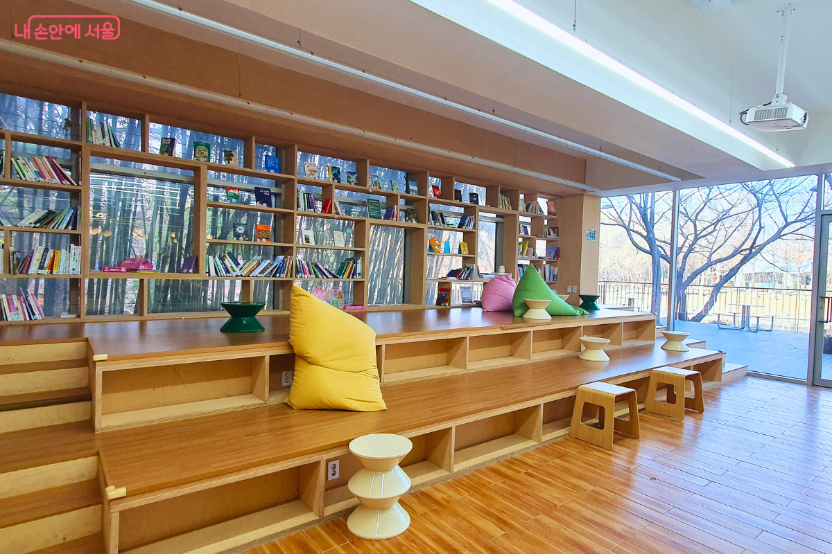 카페 한쪽 벽면을 채운 계단식 도서 공간이다. 원목으로 짜인 넓은 계단형 좌석 위에 노란색, 분홍색, 초록색의 큰 빈백 소파가 놓여 있으며, 벽면 책장에는 다양한 도서들이 비치되어 있다. 어린이와 가족 방문객이 편안하게 앉아 책을 읽으며 쉴 수 있는 도서관 형태의 휴식 공간으로 조성되었다.