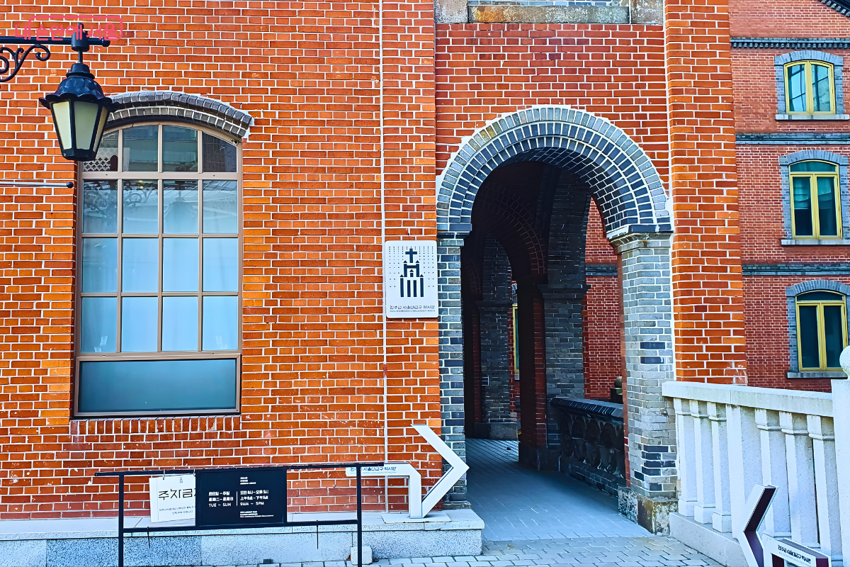 사도회관 건물의 입구와 측면 일부를 담은 외부 사진이다. 왼쪽 벽면에는 격자형 창문과 금속제 가스등 양식 조명이 매달려 있다. 벽면 중앙에는 '천주교 서울대교구 역사관 사도회관'이라고 적힌 흰색 표지판이 있으며, 오른쪽에는 아치형 통로가 배치되어 건물 내부와 연결된다. &copy;양정화