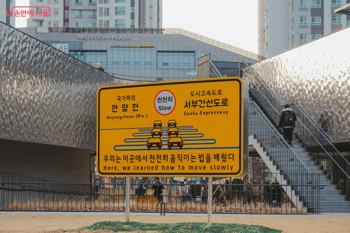 노란색 교통표지판 형태의 설치 작품. '천천히 Slow' 마크와 함께 '우리는 이곳에서 천천히 움직이는 법을 배웠다'라는 글귀가 적혀 있다.
