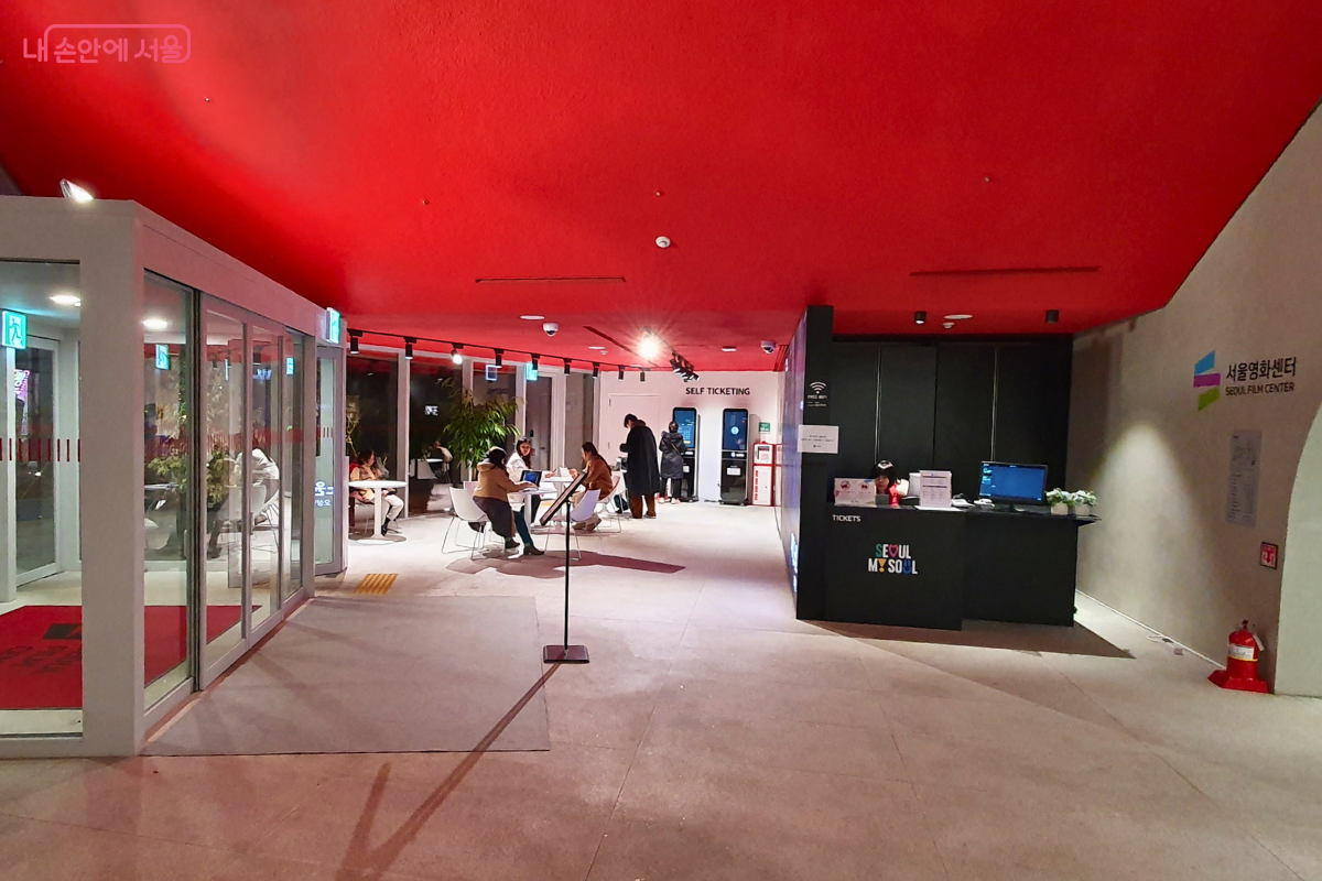 서울영화센터 1층 로비의 내부 모습이다. 천장은 강렬한 붉은색으로 마감되어 있으며, 오른쪽에는 'SEOUL MY SOUL' 로고가 새겨진 검은색 안내 데스크가 위치한다. 중앙과 왼쪽에는 방문객들이 앉아서 쉴 수 있는 하얀색 테이블과 의자들이 배치되어 있고, 배경에는 무인 발권기가 보인다. &copy;신창근