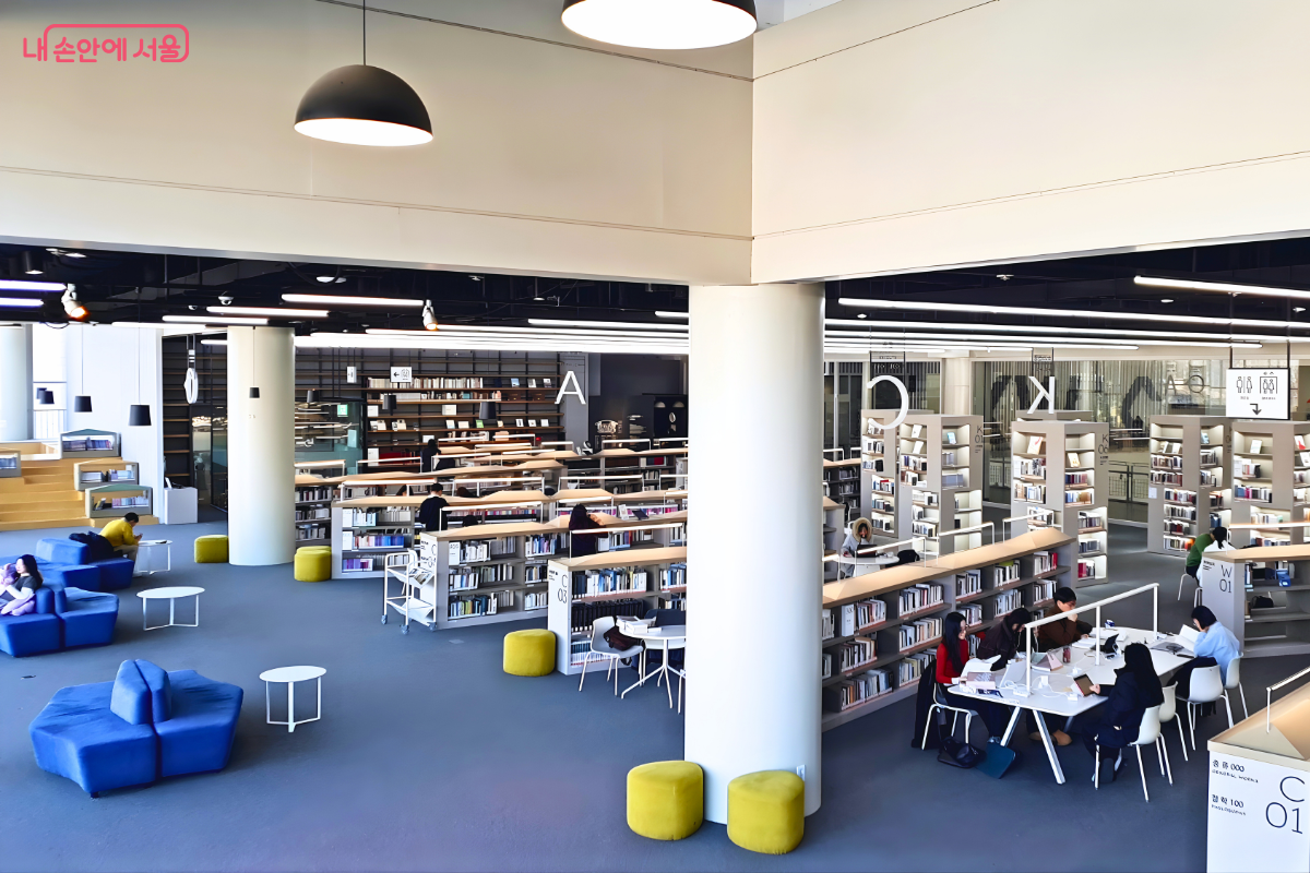 도서관 내부의 전체적인 열람 공간 모습이다. 왼쪽에는 파란색의 낮은 소파들이 배치되어 있고, 중앙에는 커다란 흰색 원형 기둥이 세워져 있다. 우측 하단 테이블에는 여러 명이 앉아 노트북 작업을 하고 있으며, 그 뒤로 A, C, K 등의 알파벳이 적힌 낮은 서가들이 질서 있게 줄지어 있다. 곳곳에 노란색 원형 스툴이 놓여 있다.