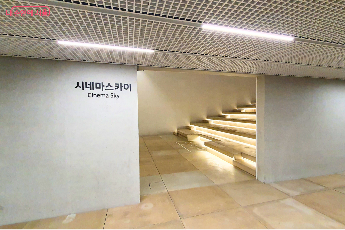 '시네마스카이 Cinema Sky'라고 적힌 벽면 뒤로 펼쳐진 야외 상영관 형태의 계단식 좌석이다. 나무 소재로 된 넓은 계단마다 아래쪽에서 은은한 간접 조명이 흘러나와 고급스러운 분위기를 연출한다. 실내와 실외가 연결되는 듯한 개방적인 구조로 설계되어 있다. &copy;신창근