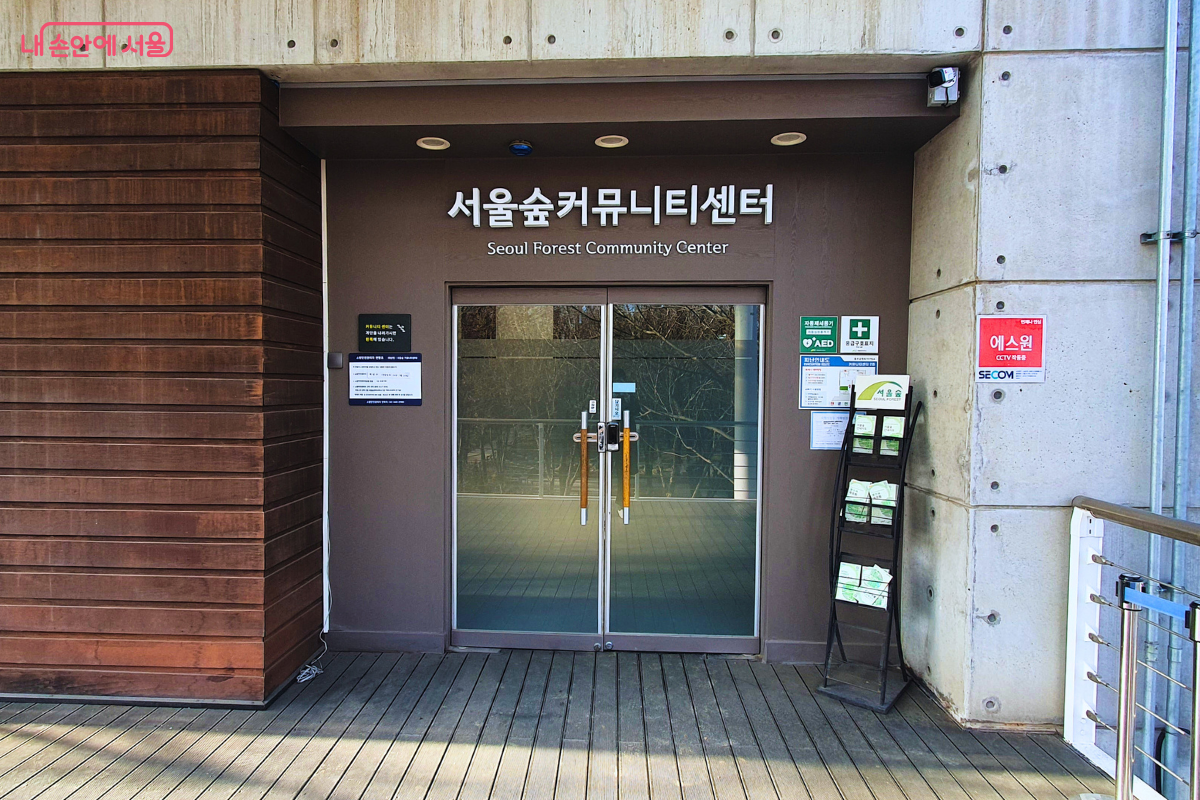 서울숲 9번 출입구 보행교와 바로 연결되는 서울숲 커뮤니티센터의 2층 진입로 모습이다. 갈색 목재 외벽과 노출 콘크리트가 조화를 이룬 건물 정면에 &lsquo;서울숲커뮤니티센터&rsquo;라는 흰색 입체 간판이 부착되어 있다. 입구 옆에는 공원 안내 리플릿 거치대와 자동심장충격기(AED) 설치 안내 표지판이 있다. &copy;양정화