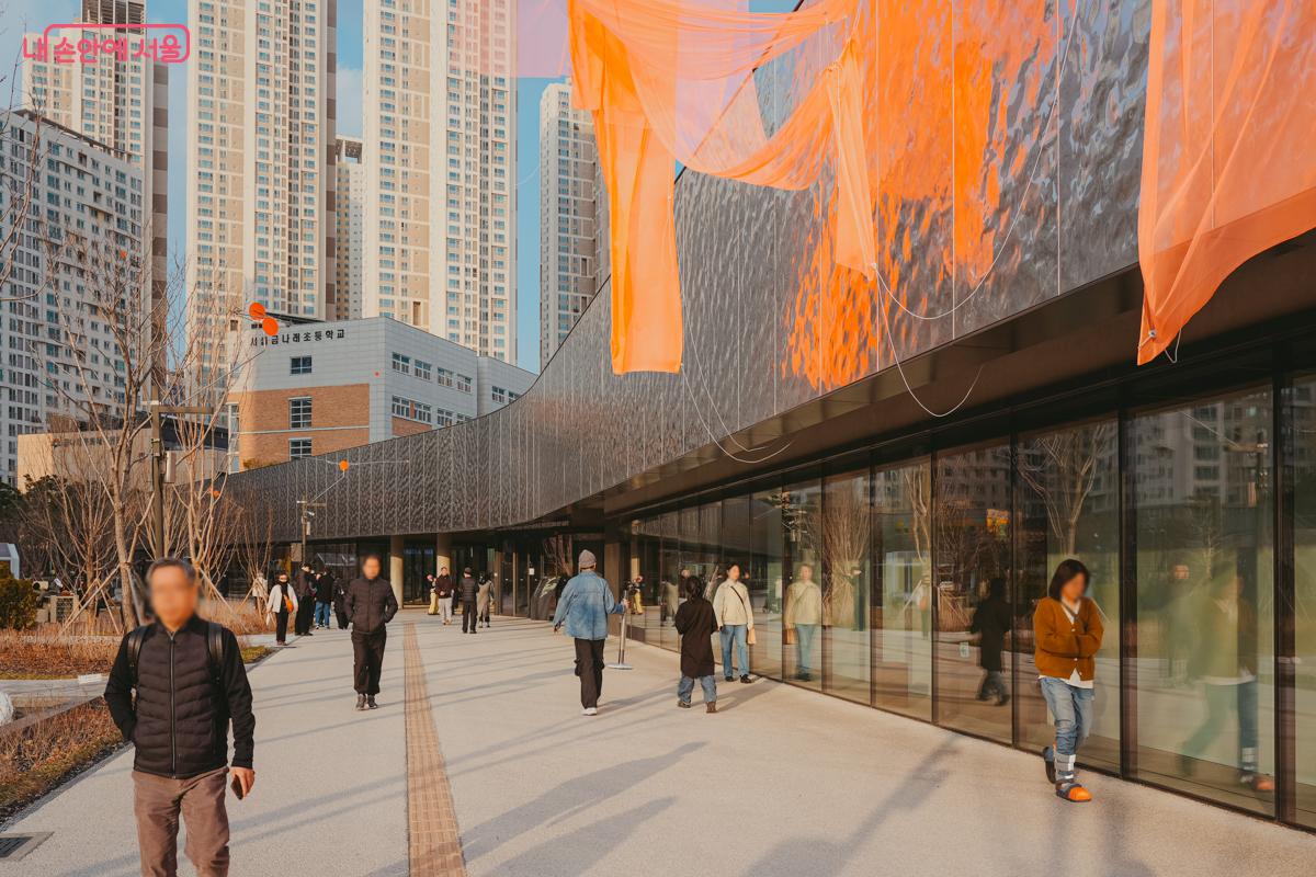 미술관 외벽에 길게 설치된 주황색 반투명 천 조형물. 바람에 흩날리는 주황색 천 아래로 시민들이 걷고 있다.