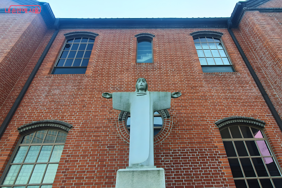 사도회관의 붉은 벽돌 외벽 중앙에 세워진 회색 석조 예수상의 정면 사진이다. 예수상은 양팔을 넓게 벌린 십자가 자세를 취하고 있으며, 그 뒤로 붉은 벽돌 벽면의 아치형 창문들이 배경을 이루고 있다. &copy;양정화