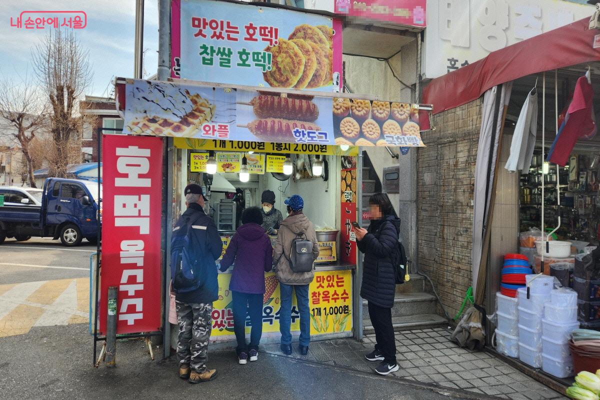 남구로시장 한 점포에서 호떡과 찐옥수수를 판매하고 있다. 판매대 앞에는 음식을 구매하려는 사람들이 줄을 서서 기다리고 있다.
