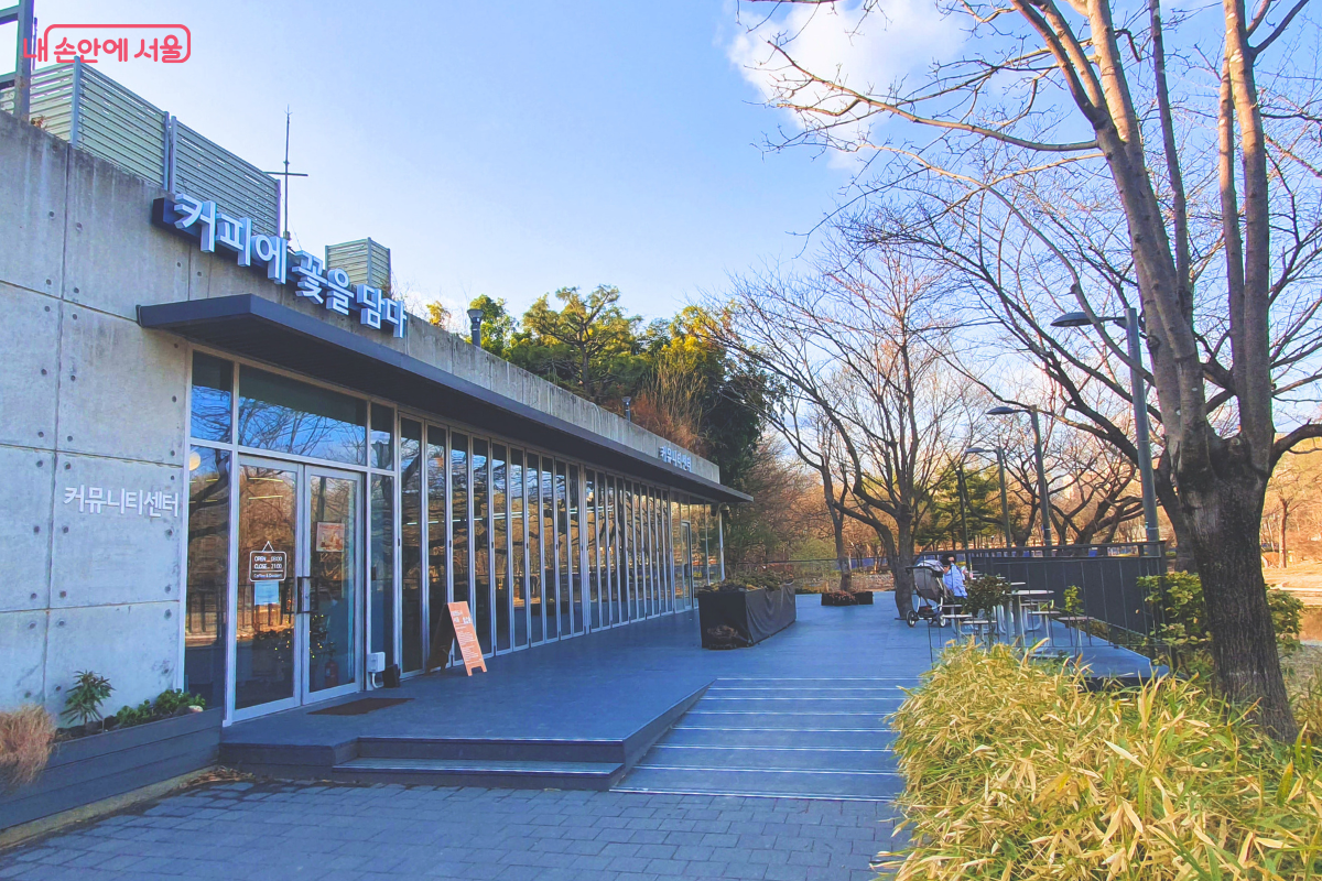 서울숲 수변 커뮤니티센터 1층에 새로 문을 연 카페 &lsquo;커피에 꽃을 담다&rsquo;의 외부 전경이다. 노출 콘크리트 공법으로 지어진 현대적인 건물 외벽에 카페 상호가 붙어 있으며, 전면은 통유리창으로 설계되어 내부가 들여다보인다. 카페 앞에는 넓은 목재 데크가 설치되어 있고 주변에 나무들이 식재되어 있어 숲속 쉼터 같은 분위기를 자아낸다. &copy;양정화