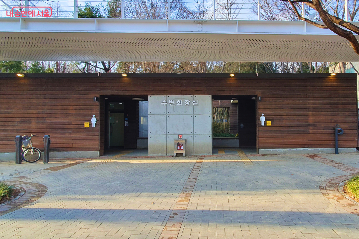 카페 건물 바로 옆에 위치한 &lsquo;수변 화장실&rsquo; 건물의 외부 전경이다. 목재와 노출 콘크리트로 마감된 단층 건물로, 중앙에 &lsquo;수변화장실&rsquo;이라는 현판이 붙어 있고 좌우로 남녀 화장실 픽토그램이 선명하게 표시되어 있다. 상단에는 보행교가 지나가는 구조이며 주변 산책로와 자연스럽게 연결되어 있다. &copy;양정화