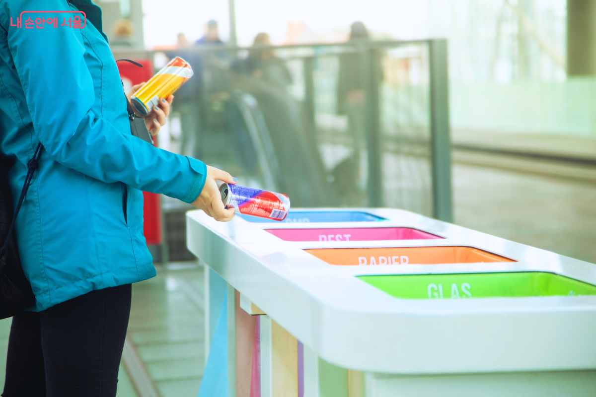 공공장소에서 푸른색 겉옷을 입은 시민이 다용도 분리수거함에 캔을 배출하려는 장면이다. 시민은 왼손에 노란색 음료 캔을 들고 있고, 오른손으로 파란색 음료 캔을 수거함 투입구 근처로 가져가고 있다. 배경으로 이동하는 사람들의 모습이 흐릿하게 보인다.