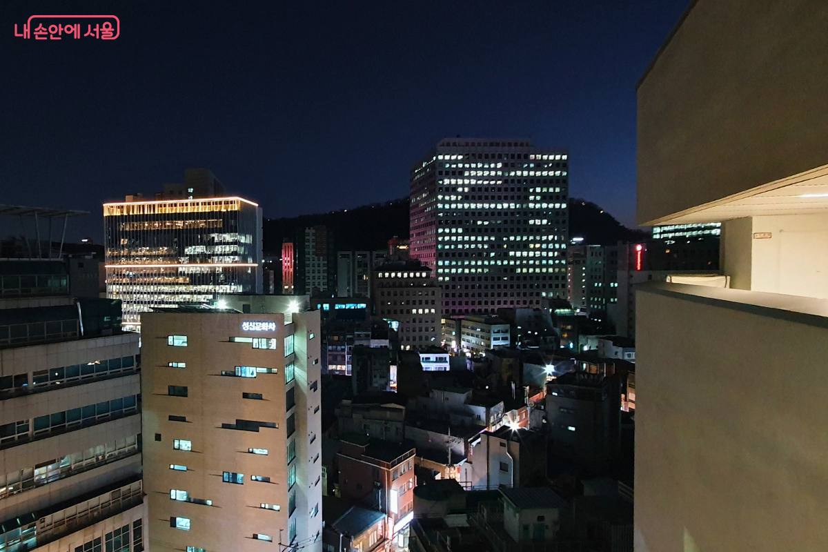 서울영화센터 옥상 층인 시네마스카이에서 바라본 서울 도심의 야경이다. 수많은 빌딩들이 각기 다른 불빛을 내뿜으며 밀집해 있고, 멀리 산의 능선이 밤하늘과 경계를 이루고 있다. 도시의 역동적인 밤 풍경이 한눈에 들어오는 탁 트인 조망을 제공한다. &copy;신창근