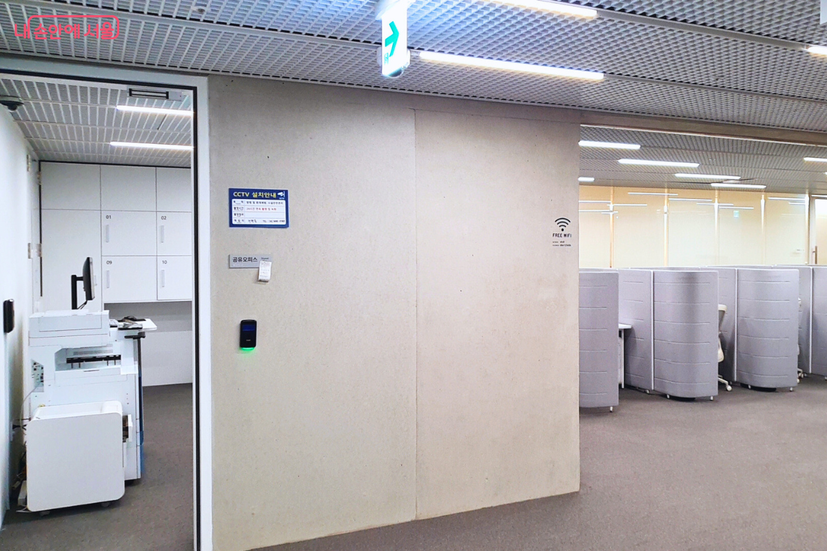 서울영화센터 내부에 마련된 공유 오피스 입구와 내부 전경이다. 왼쪽에는 복합기와 사물함이 구비되어 있으며, 오른쪽 개방된 공간에는 회색 파티션으로 구분된 개인 업무용 책상들이 나란히 배치되어 있다. 벽면에는 '공유오피스' 명칭과 함께 무료 와이파이 안내 및 CCTV 설치 안내문이 부착되어 있다. &copy;신창근