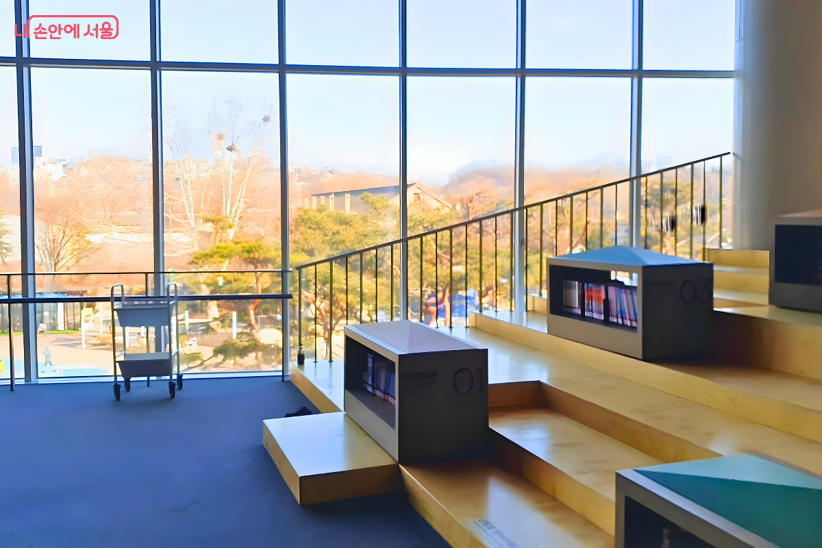 대형 통창을 배경으로 설치된 밝은 나무 재질의 계단식 열람석이다. 계단 곳곳에는 '01', '03' 등의 번호가 적힌 회색 박스 형태의 낮은 서가가 놓여 있으며 그 안에 책들이 들어 있다. 계단 좌측 하단 바닥에는 '신발을 벗고 이용해 주세요'라는 문구가 부착되어 있다. 창밖으로는 나무들이 있는 야외 풍경이 넓게 펼쳐져 있다.