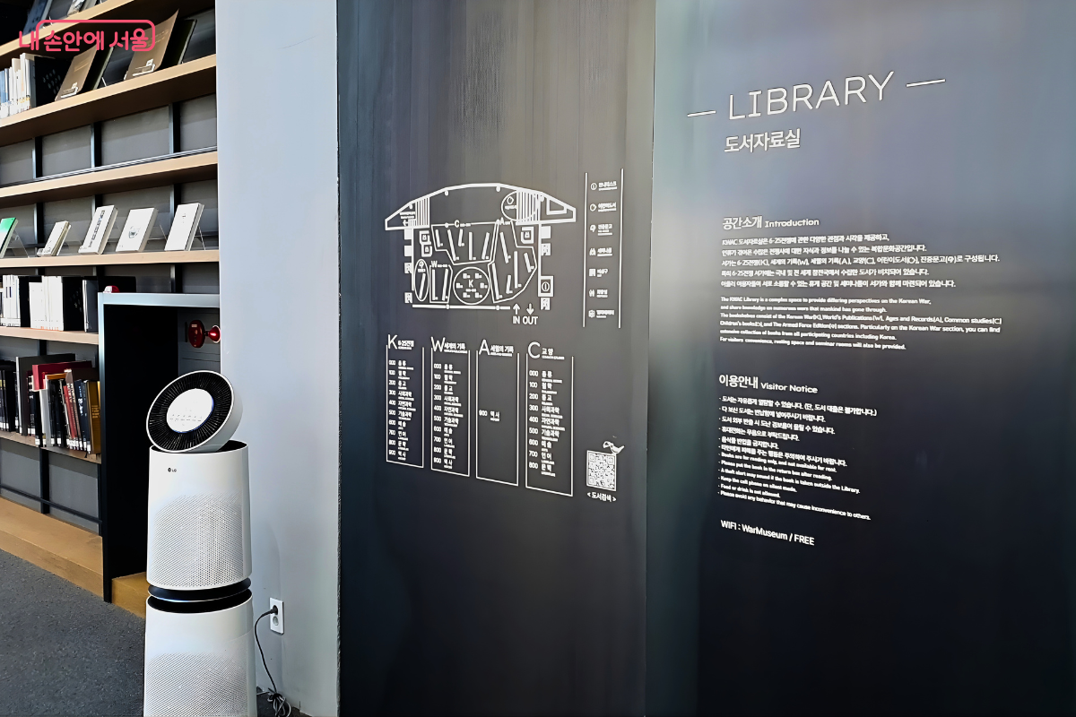 도서관 내부의 검은색 벽면에 부착된 안내 정보이다. 벽면에는 도서관의 평면도와 함께 K(6&middot;25전쟁), W(세계의 기록), A(세상의 기록), C(교양) 등의 분류 코드가 상세히 적혀 있다. 오른쪽에는 'LIBRARY 도서자료실'이라는 제목 아래 '공간소개'와 '이용안내' 문구가 국문과 영문으로 기재되어 있다. 좌측 하단에는 흰색 공기청정기가 놓여 있고 그 뒤로 책들이 꽂힌 서가가 보인다.