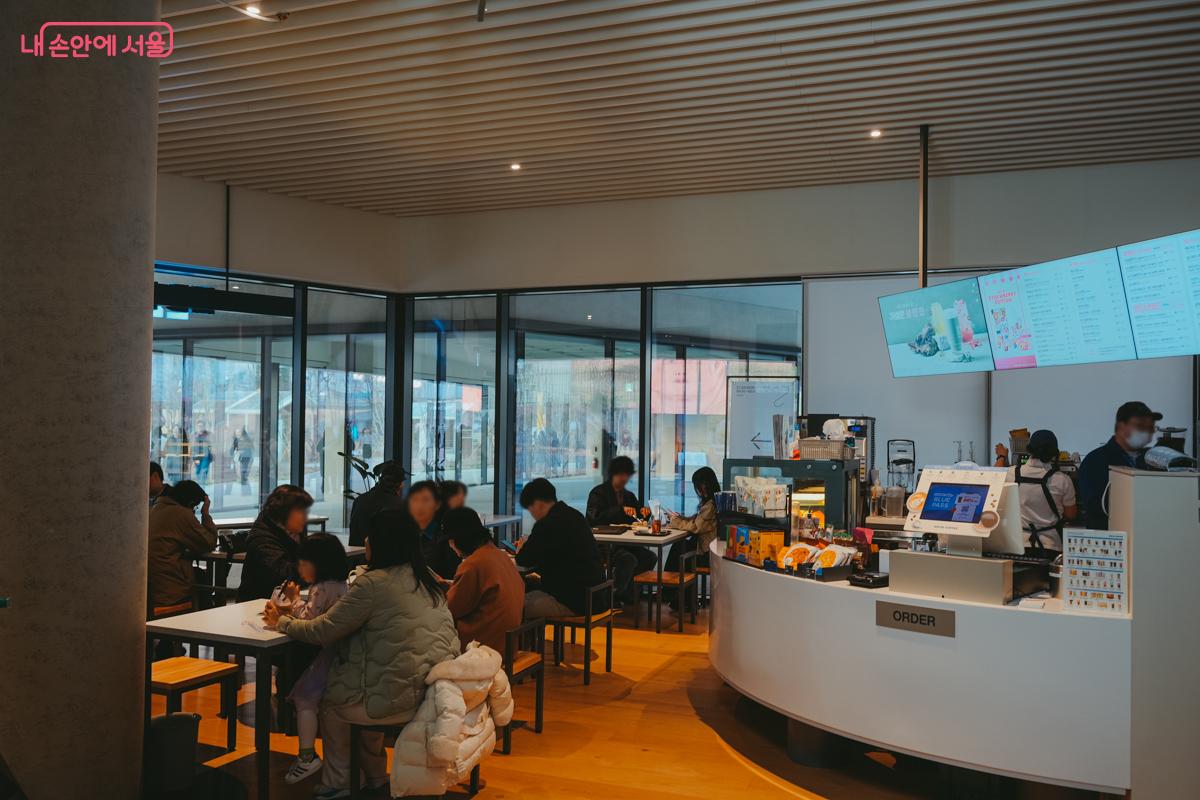 통유리창으로 된 미술관 내 카페테리아. 테이블에 앉아 커피를 마시며 대화를 나누는 시민들로 북적인다.