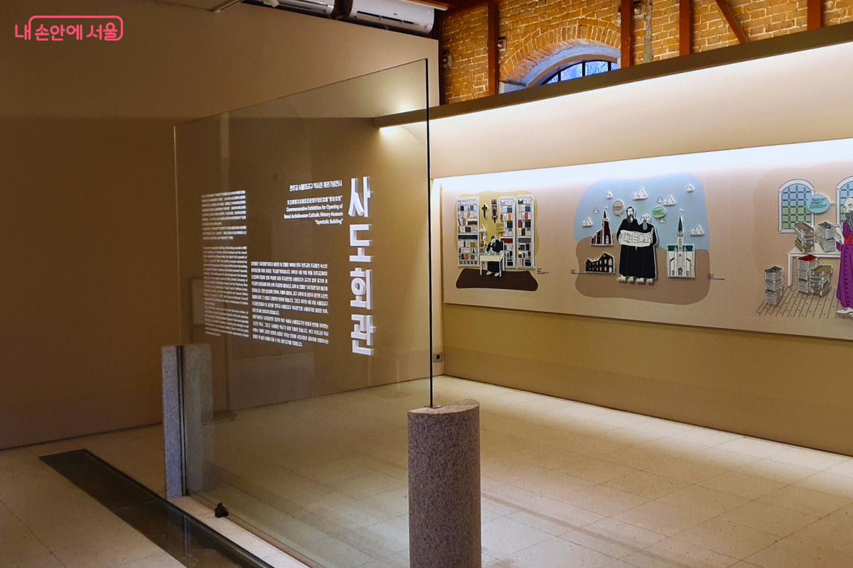 사도회관 내부 전시실 모습이다. 전면에 '사도회관' 명칭과 안내 문구가 적힌 대형 유리 판이 세워져 있으며, 그 뒤로 사제들의 모습과 건물을 그린 일러스트 벽화가 보인다. 바닥은 밝은색 타일이며 천장에는 전시용 트랙 조명이 설치되어 있다. &copy;양정화