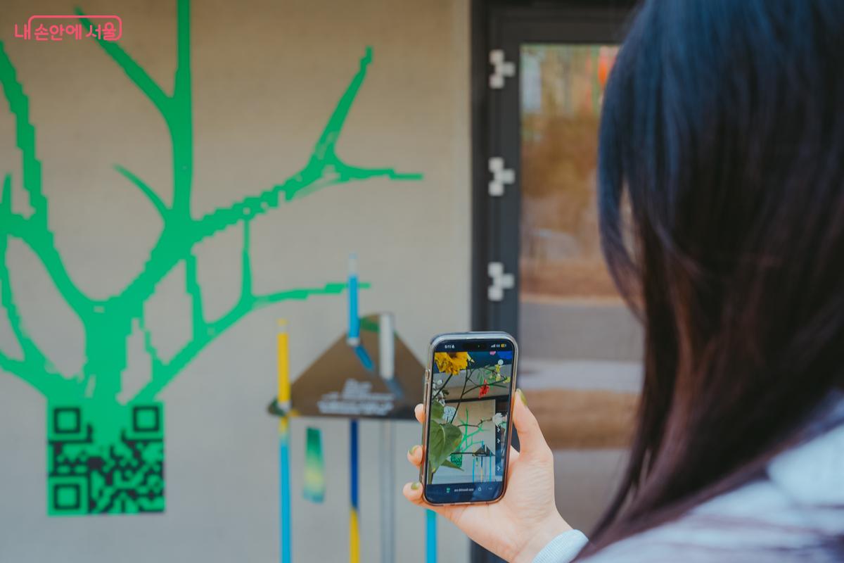 한 관람객이 스마트폰 카메라로 벽면에 그려진 초록색 나무 모양의 QR코드를 비추며 증강현실(AR) 작품을 체험하고 있다.